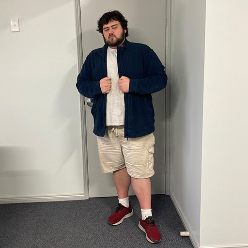 The image shows a man standing in an office hallway. He is wearing a dark blue zip-up jacket, a white t-shirt, beige cargo shorts, and red sneakers with white socks. His arms are slightly bent, and his legs are apart, giving him a relaxed posture. The background features a plain white wall and a gray carpeted floor, with a door visible behind him. The man has curly hair and a beard, and he appears to be casually dressed.